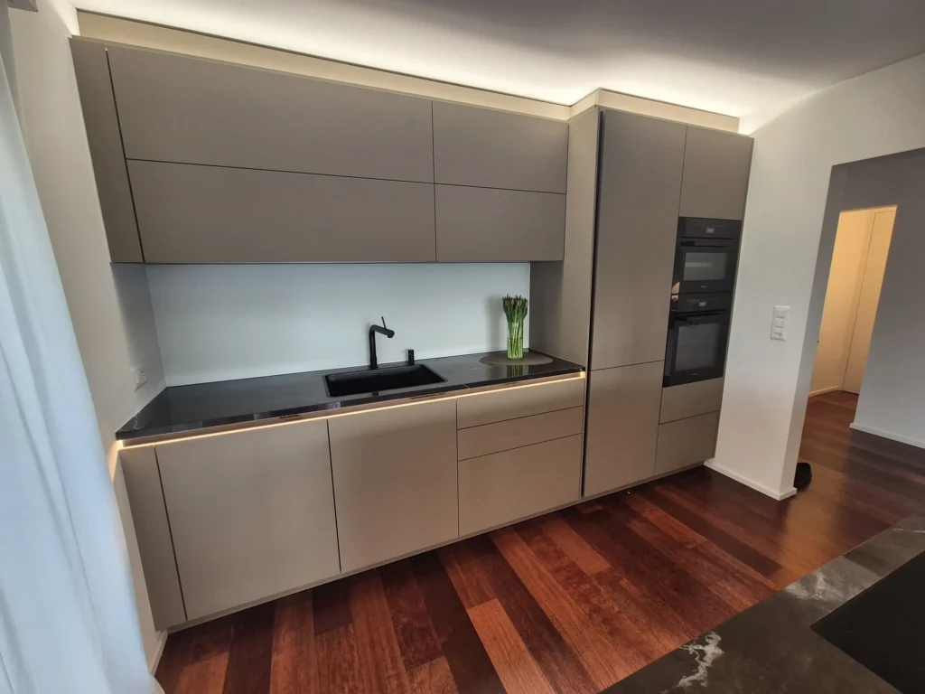 Küchenzeile mit beigegrauen Fronten, dunkler Arbeitsplatte, Spüle und zwei Backöfen auf rotbraunem Holzboden